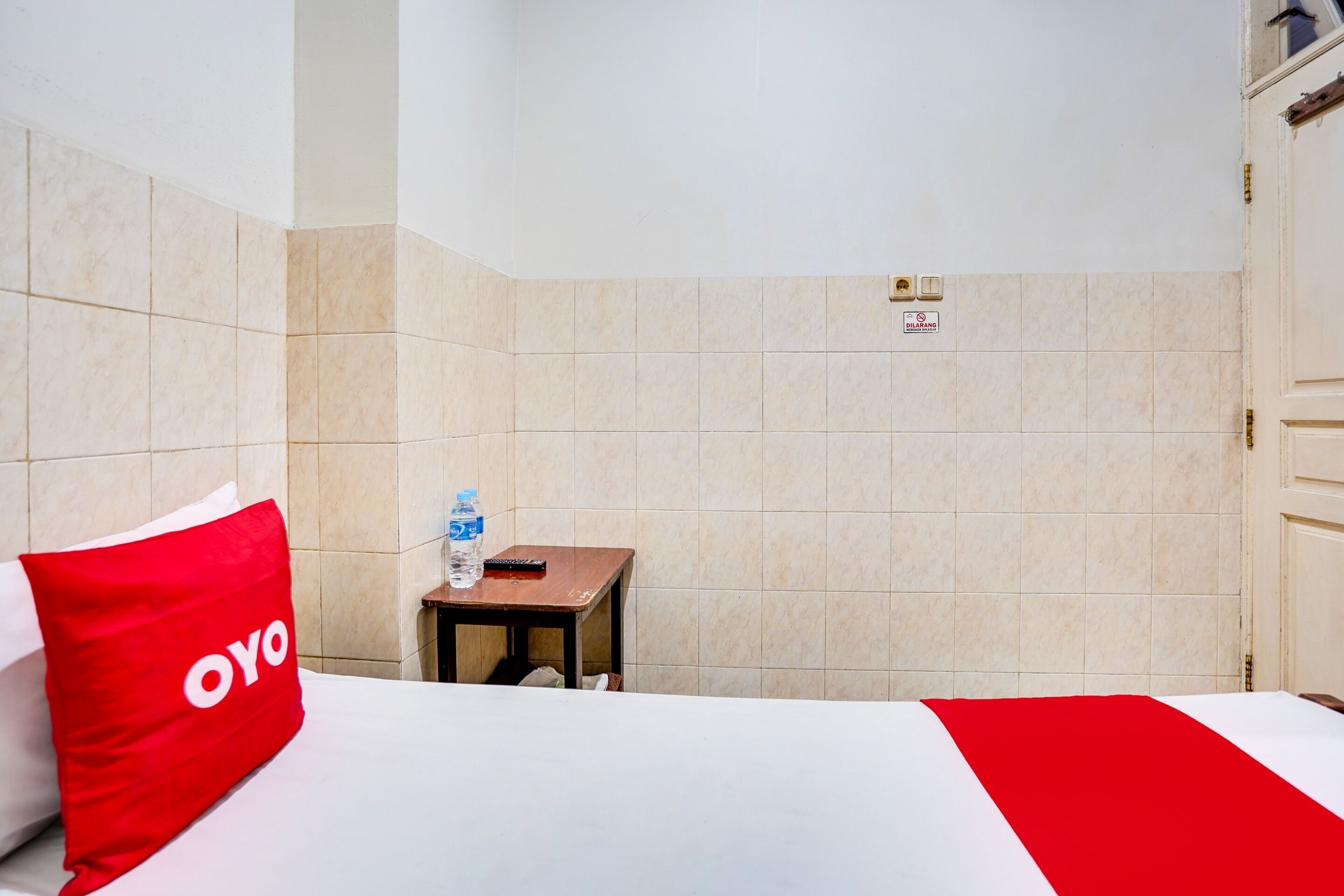 A hotel room with a bed, a red pillow, a small table with a water bottle, and a sign on the wall.