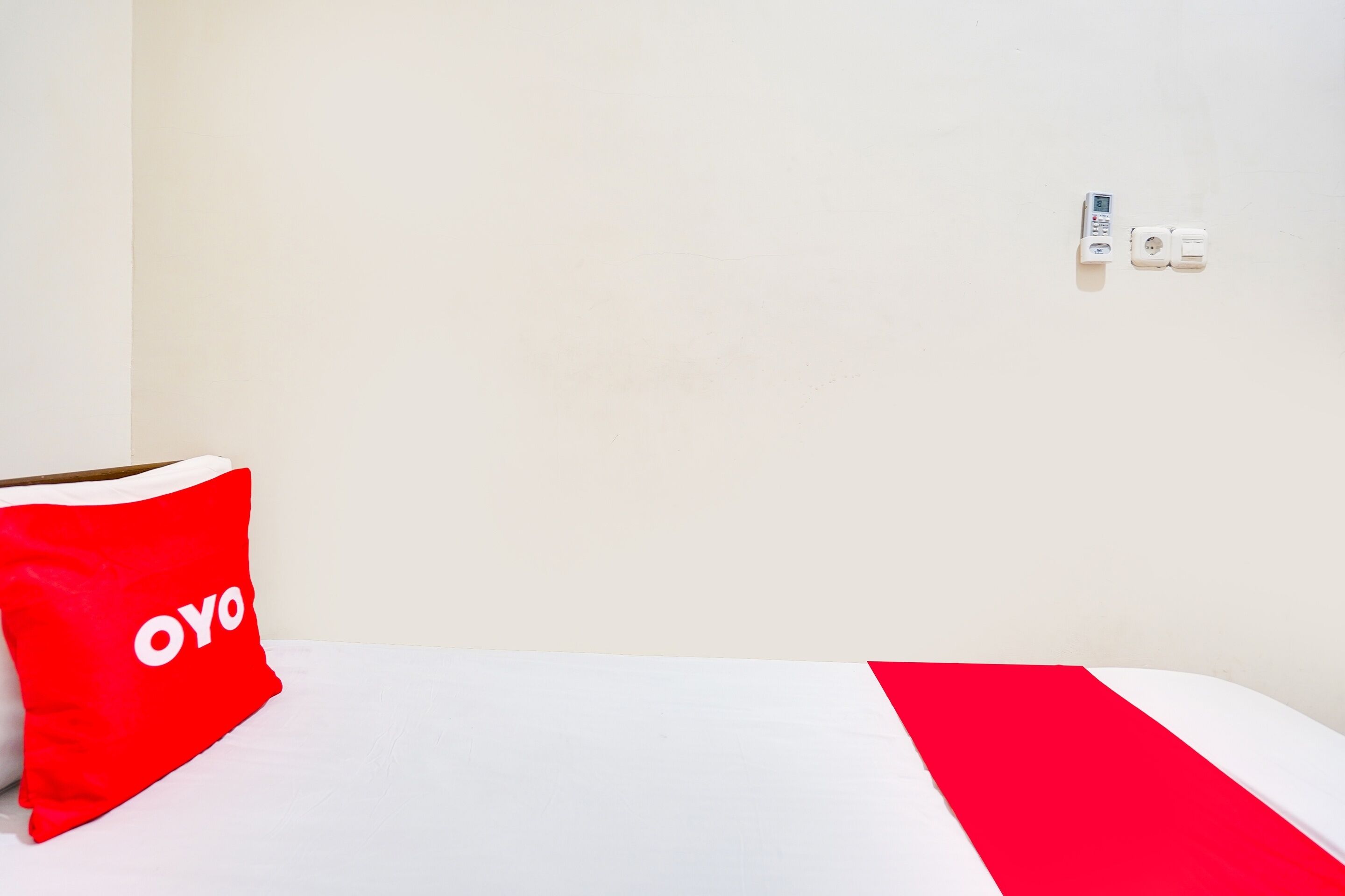 A hotel room with a white bed, a red pillow with "OYO" text, and a power outlet on the wall.
