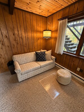 A cozy room with a wooden ceiling, a cushioned sofa with patterned cushions, a small ottoman, and a window with curtains. - Vacation Home in Quiet Wooded Setting with Beautiful Scenic Views from Living (Bessemer)