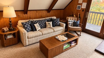 A cozy living room with a sofa, coffee table, and a chair. There is a lamp on a side table, and a door leading outside.