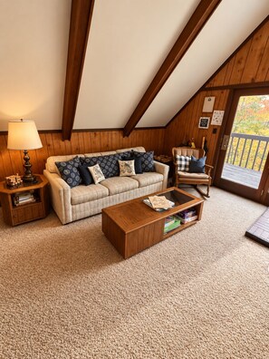A cozy living room with a sofa, coffee table, and a chair. There is a lamp on a side table, and a door leading outside.