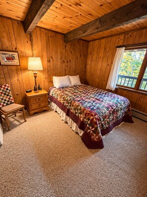 A cozy bedroom with a wooden ceiling, a bed with a colorful quilt, a nightstand with a lamp, a chair, and a window with a view of trees. - Vacation Home in Quiet Wooded Setting with Beautiful Scenic Views from Living (Bessemer)