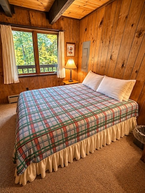 A wooden cabin bedroom with a plaid bedspread, a nightstand with a lamp, and a window with curtains. - Vacation Home in Quiet Wooded Setting with Beautiful Scenic Views from Living (Bessemer)