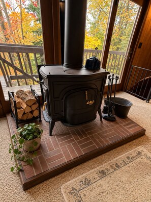 A wood-burning stove with a chimney, a log storage rack, a potted plant, and a rug on a wooden deck. - Vacation Home in Quiet Wooded Setting with Beautiful Scenic Views from Living (Bessemer)
