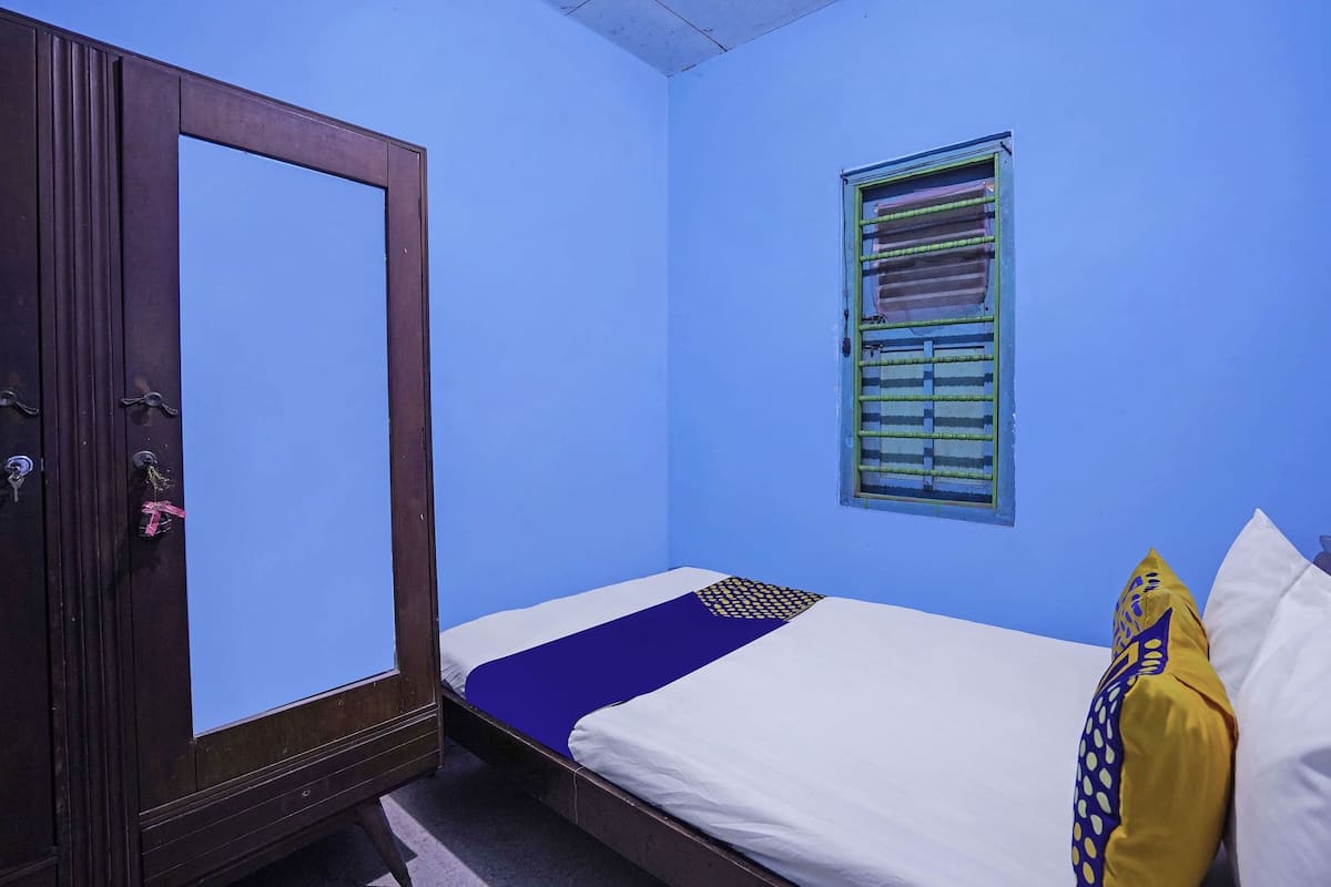 A room with a single bed, a wooden wardrobe, a window with blinds, and a small window with a curtain.