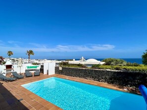 Una piscina con sillas de descanso, un muro de piedra y vistas al océano y edificios blancos.