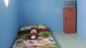 Une pièce aux murs bleus, avec un lit recouvert d’une couverture aux couleurs vives, un ventilateur de plafond et une petite armoire contenant un téléviseur.