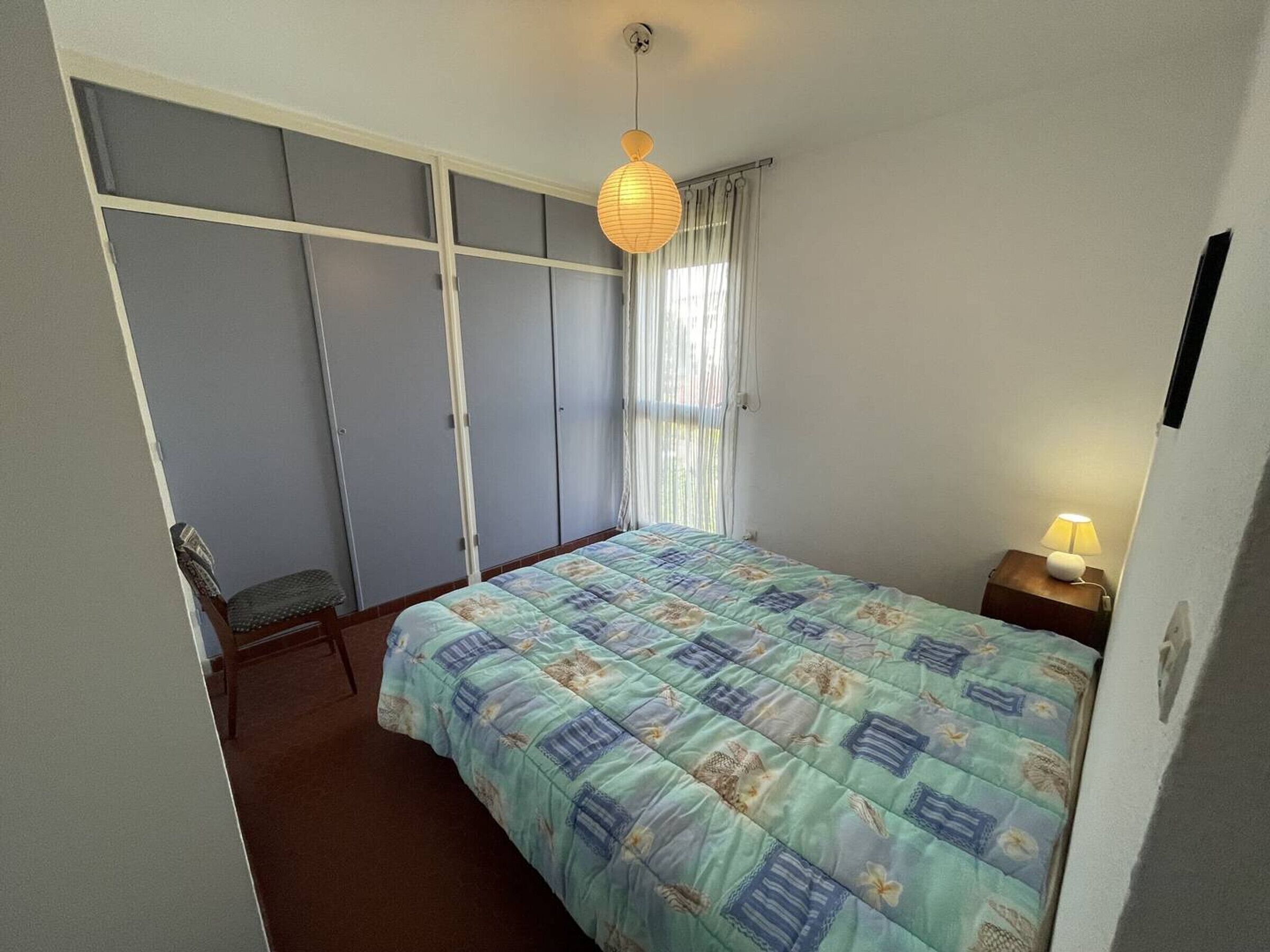 Een slaapkamer met een bed, nachtkastje, lamp, kledingkast en een raam met gordijnen.