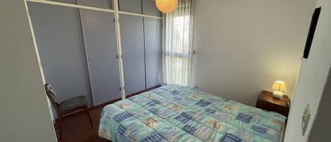Een slaapkamer met een bed, nachtkastje, lamp, kledingkast en een raam met gordijnen.