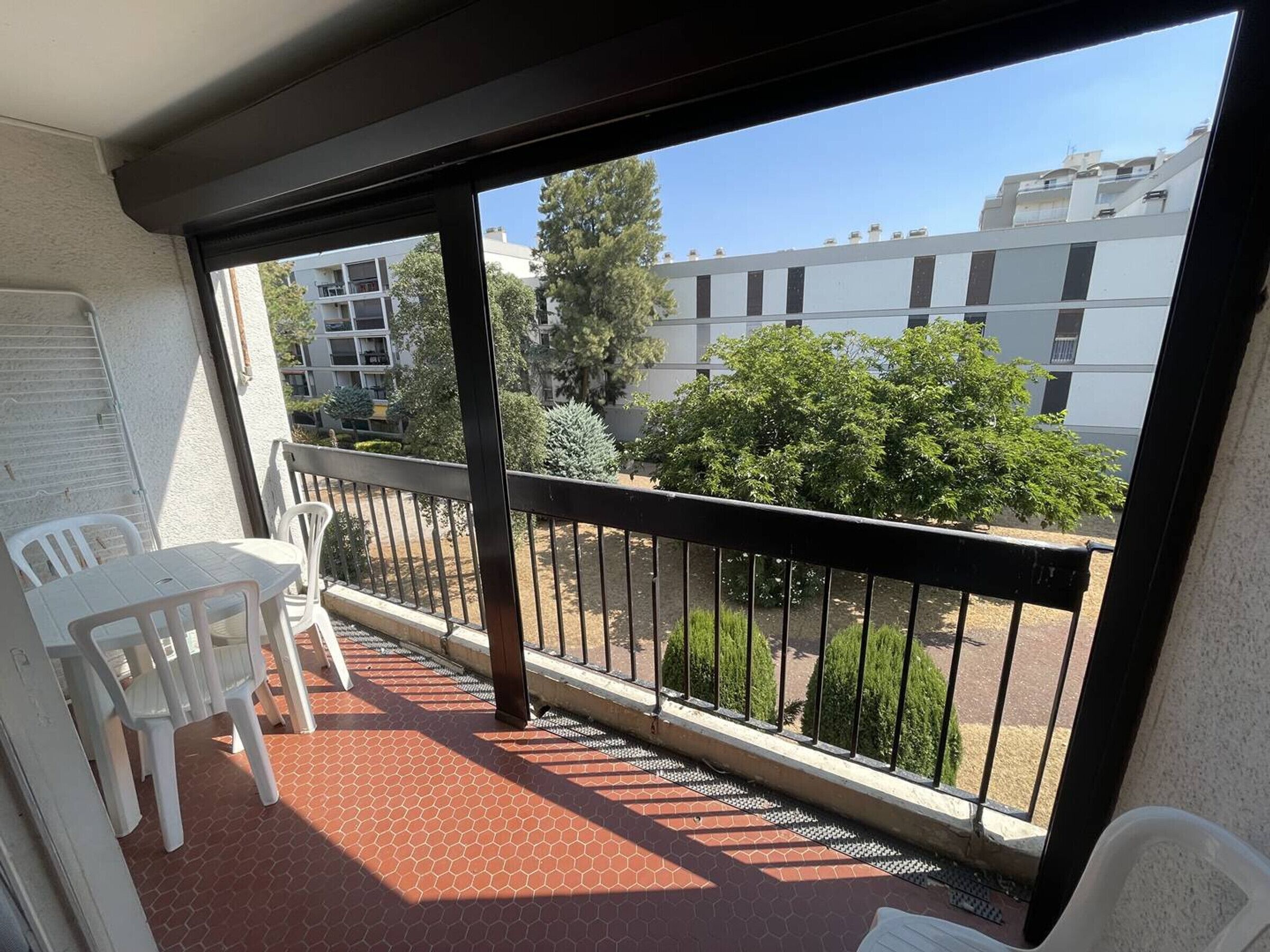 A balcony with a tiled floor, white plastic chairs, and a view of buildings and trees.