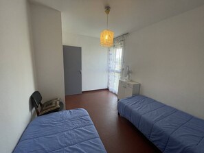 A room with two beds, a desk, a chair, a window with curtains, and a hanging light fixture. - Spacious 2-BR Apt in Saint-Cyprien, 400m to Beach, Pets Allowed (Saint-Cyprien)