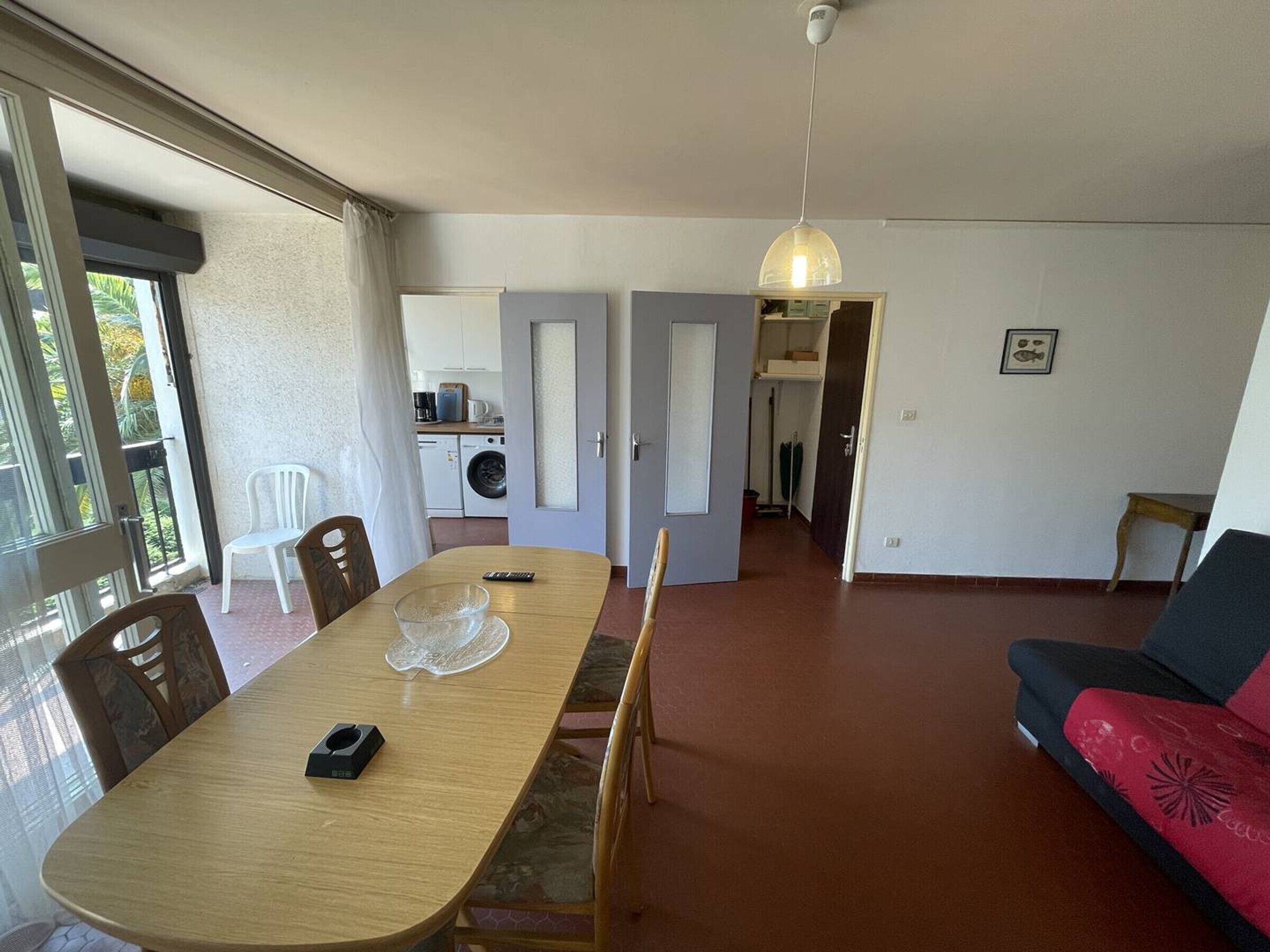 A dining area with a table, chairs, and a remote control. There is a kitchen in the background with appliances and a dining area with a sofa.