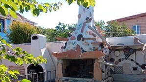 A stone-built outdoor kitchen with a chimney, surrounded by plants and a fence. - Holidays in Sardinia, apartment Arbatax (Arbatax)