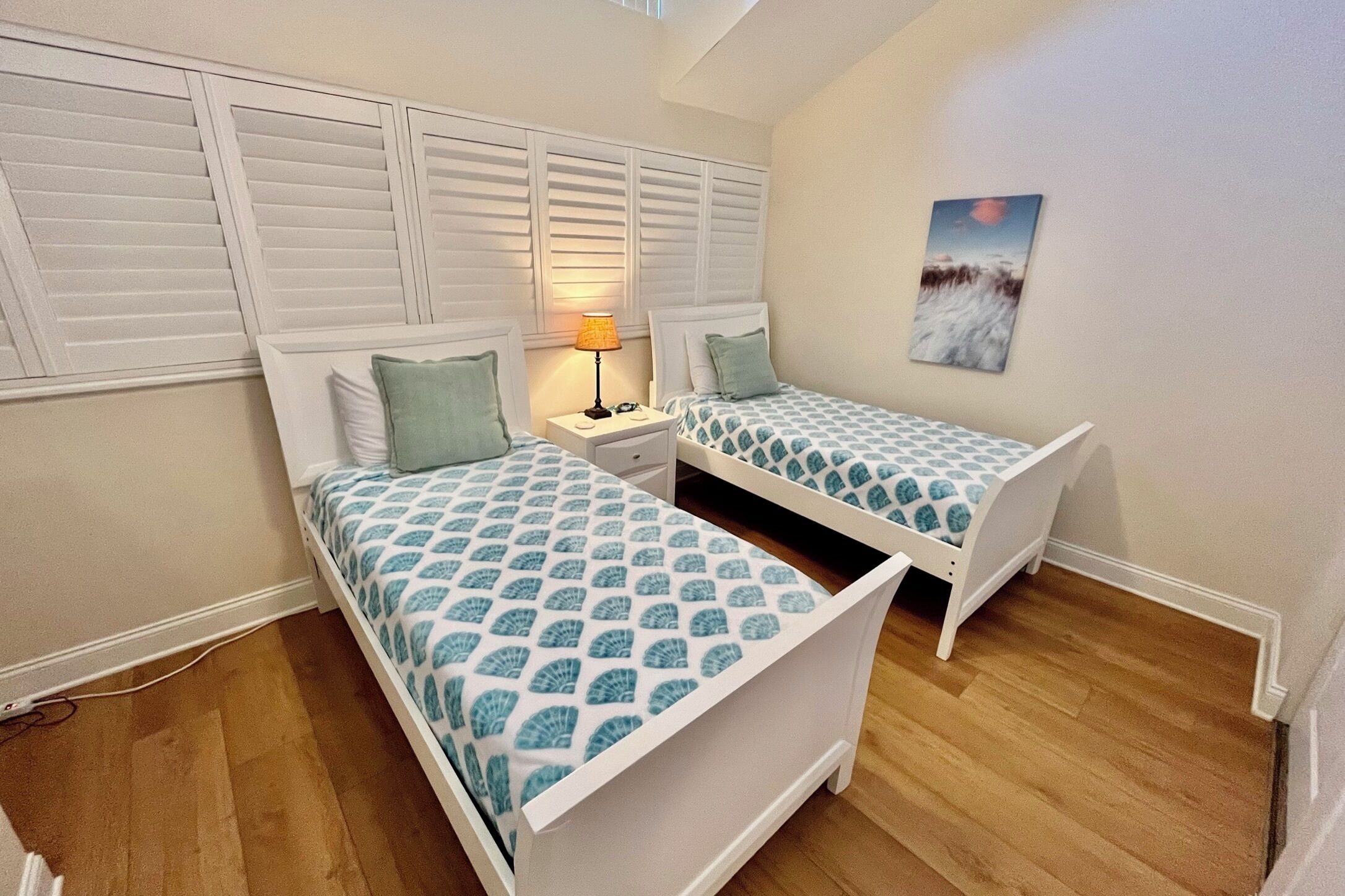 Un dormitorio con dos camas individuales, una mesita de noche con lámpara, un cuadro en la pared y techo inclinado.