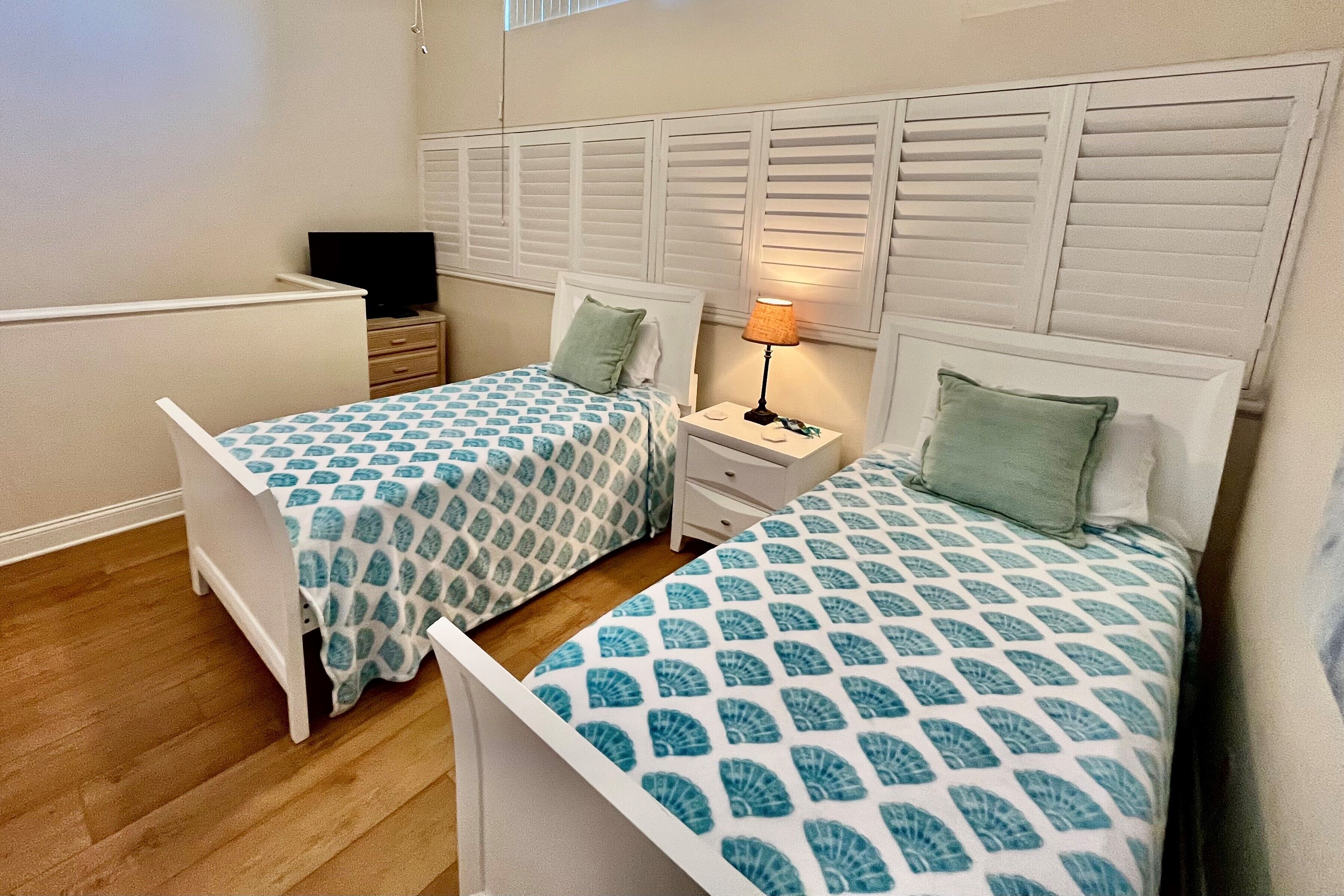 A bedroom with two single beds, a nightstand with a lamp, a TV, and a window with blinds.