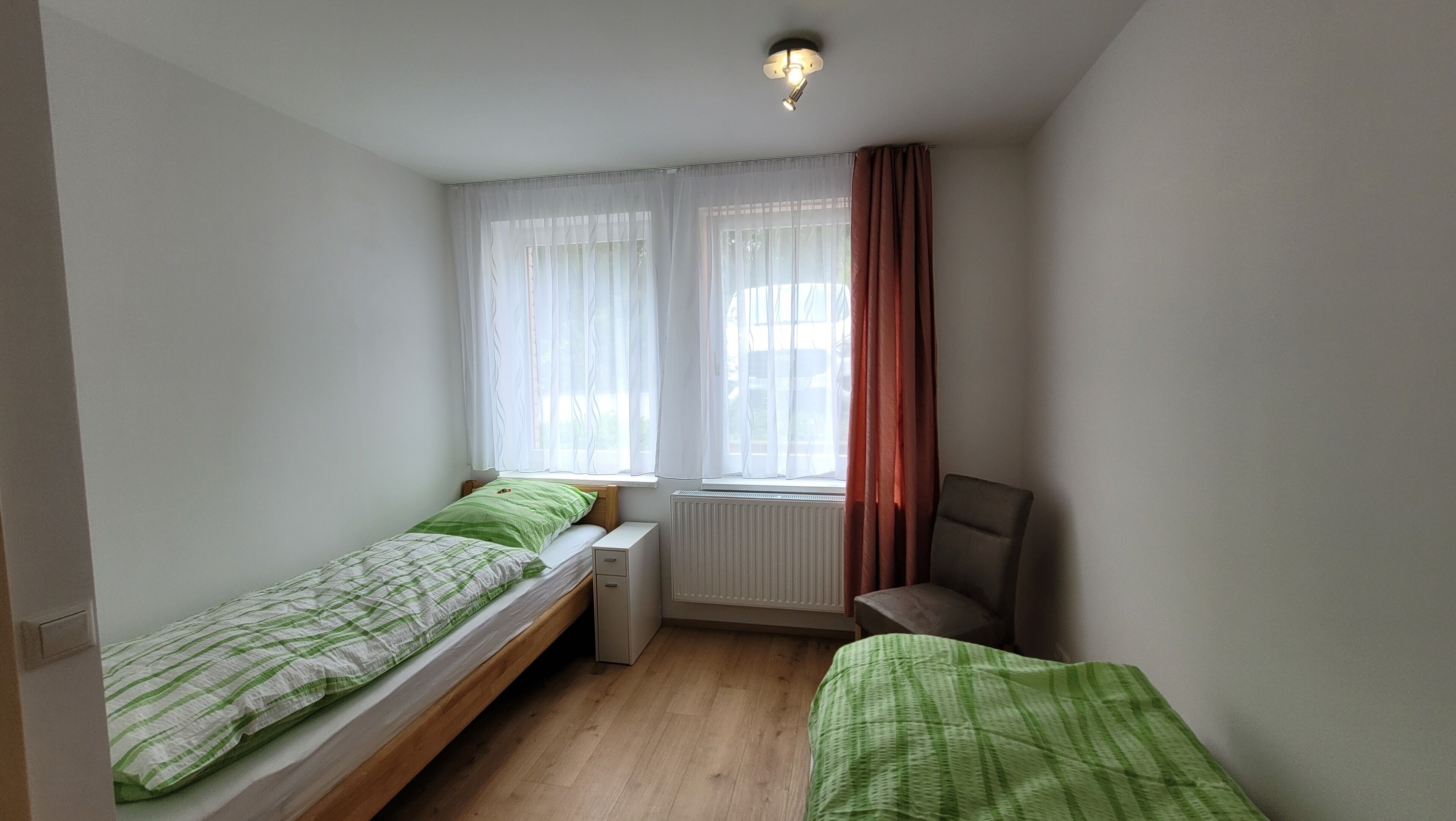 Ein kleines, gut beleuchtetes Zimmer mit zwei Einzelbetten, einem Sessel, einem Heizkörper und einem Fenster mit durchsichtigen Vorhängen.
