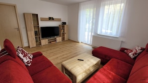 A living room with a large flat-screen TV, wooden furniture, and a window with sheer curtains.