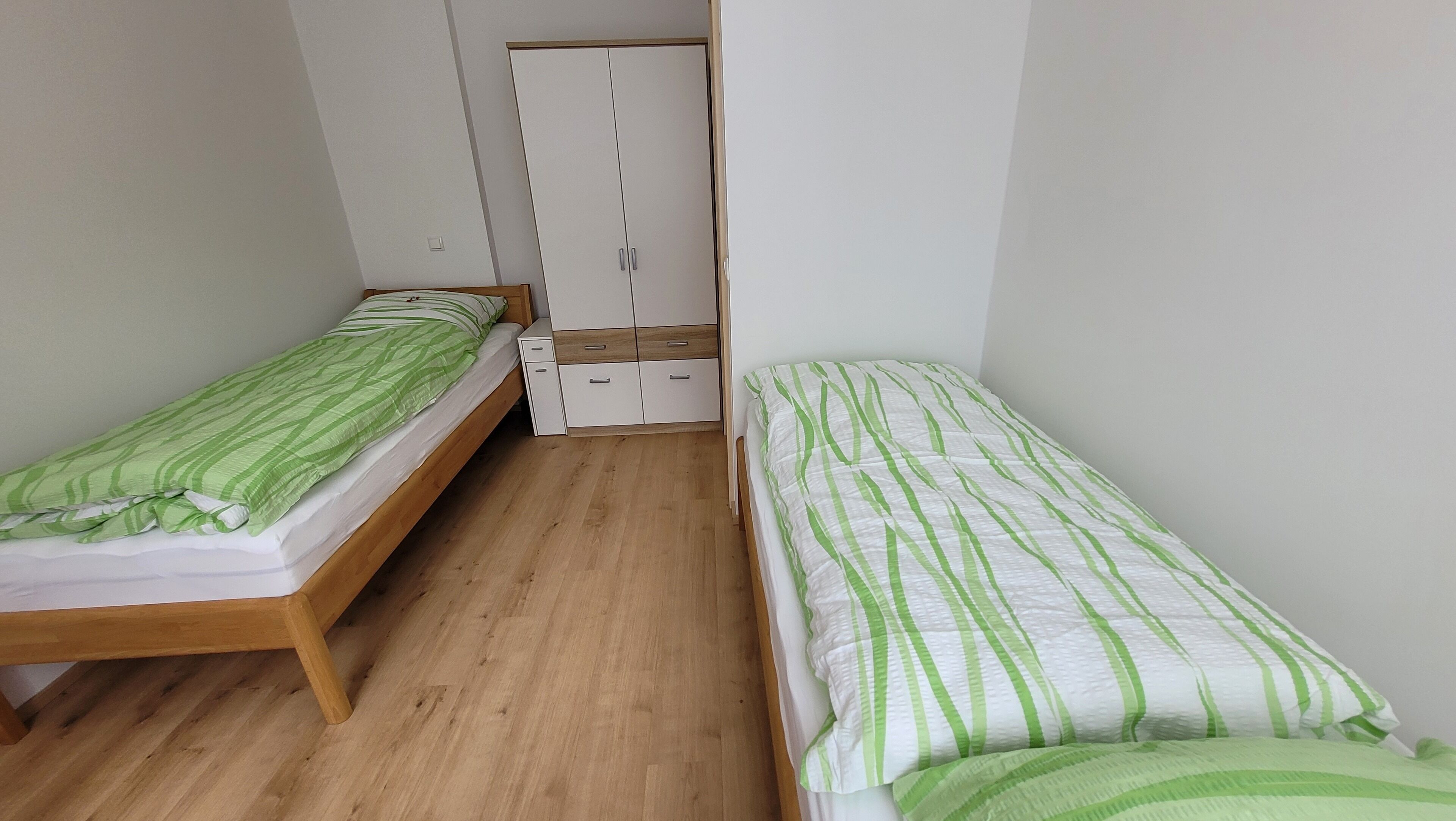 A small, narrow room with two single beds, each with green-striped bedding. The room has a wooden floor and a white wardrobe with multiple compartments.
