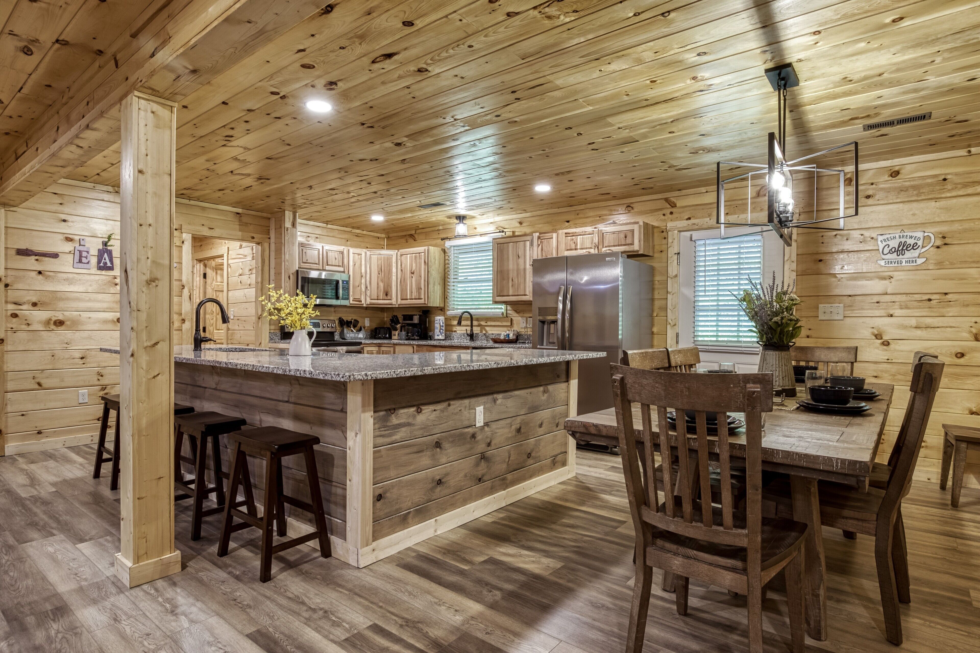 A wooden cabin interior with a kitchen featuring a central island, bar stools, wooden dining table, and chairs.
