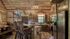 A rustic kitchen with wooden walls, a central island with stools, a stainless steel refrigerator, and a 'WELCOME' sign.