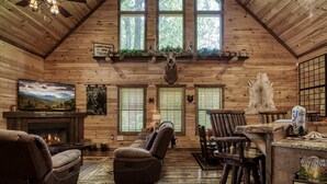 A log cabin interior with a fireplace, leather recliners, a bar area, and a mounted deer head.