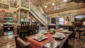 A rustic cabin interior with a wooden staircase, a dining area with a red tablecloth, and a mounted deer head.