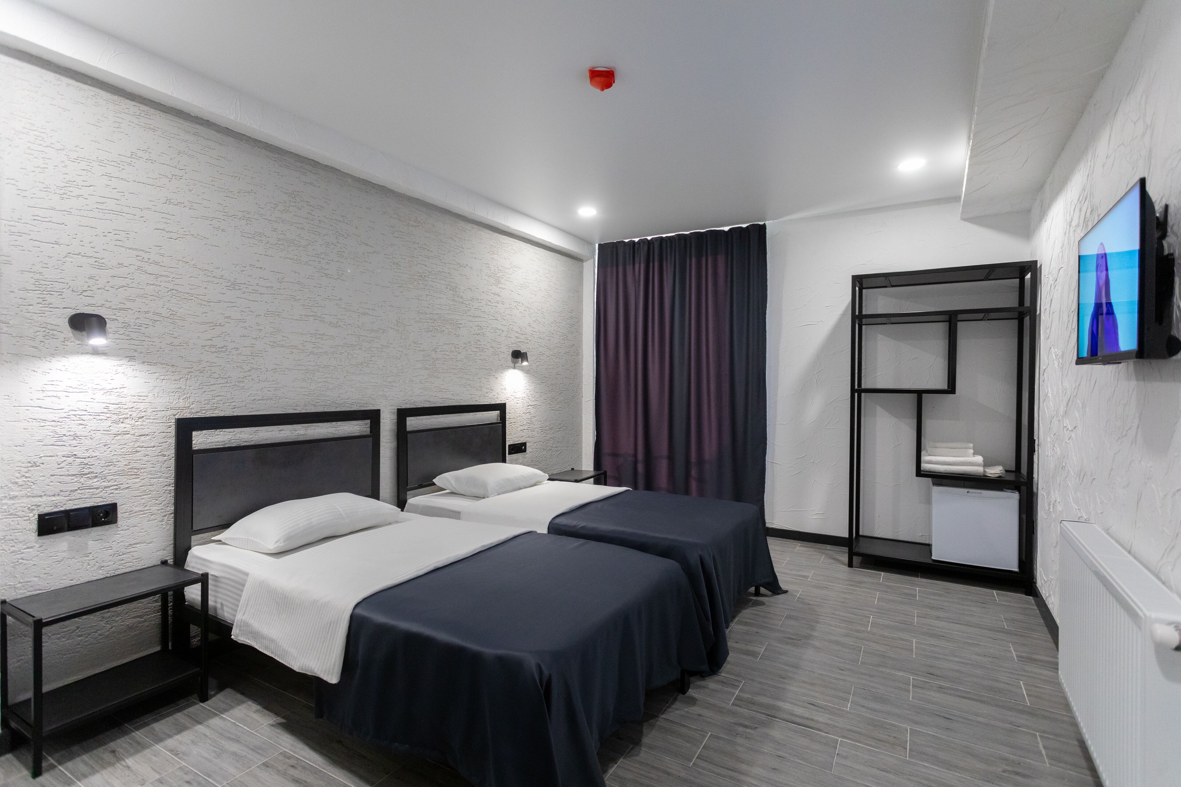 A hotel room with two beds, a TV mounted on the wall, a small shelf with towels, and a window with curtains.