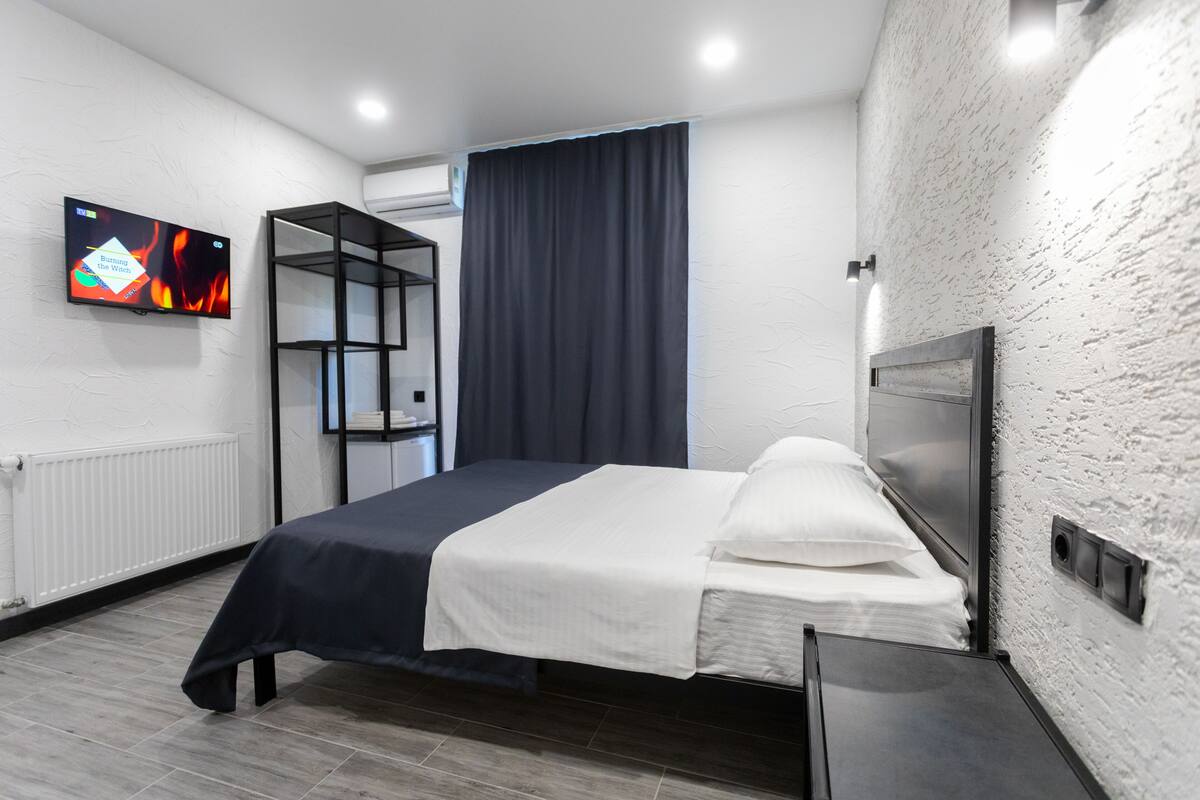 Une chambre d’hôtel moderne équipée d’un lit, d’un téléviseur fixé au mur, d’une étagère intégrée et d’une fenêtre avec des rideaux.