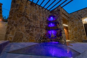 Una fuente de piedra con niveles iluminados y una piscina cristalina al frente.