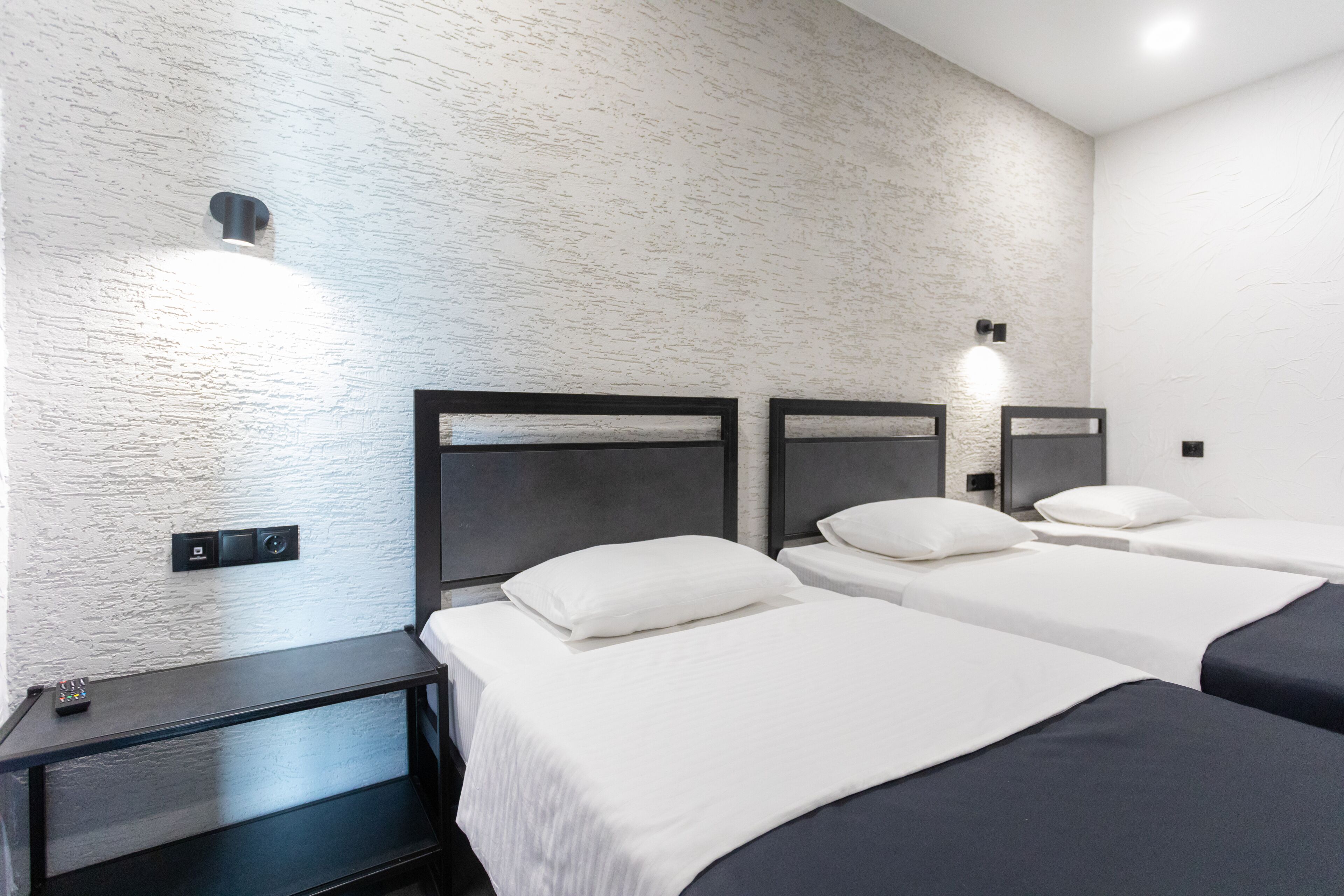A hotel room with three single beds, a small desk with a remote control, and a textured white wall.