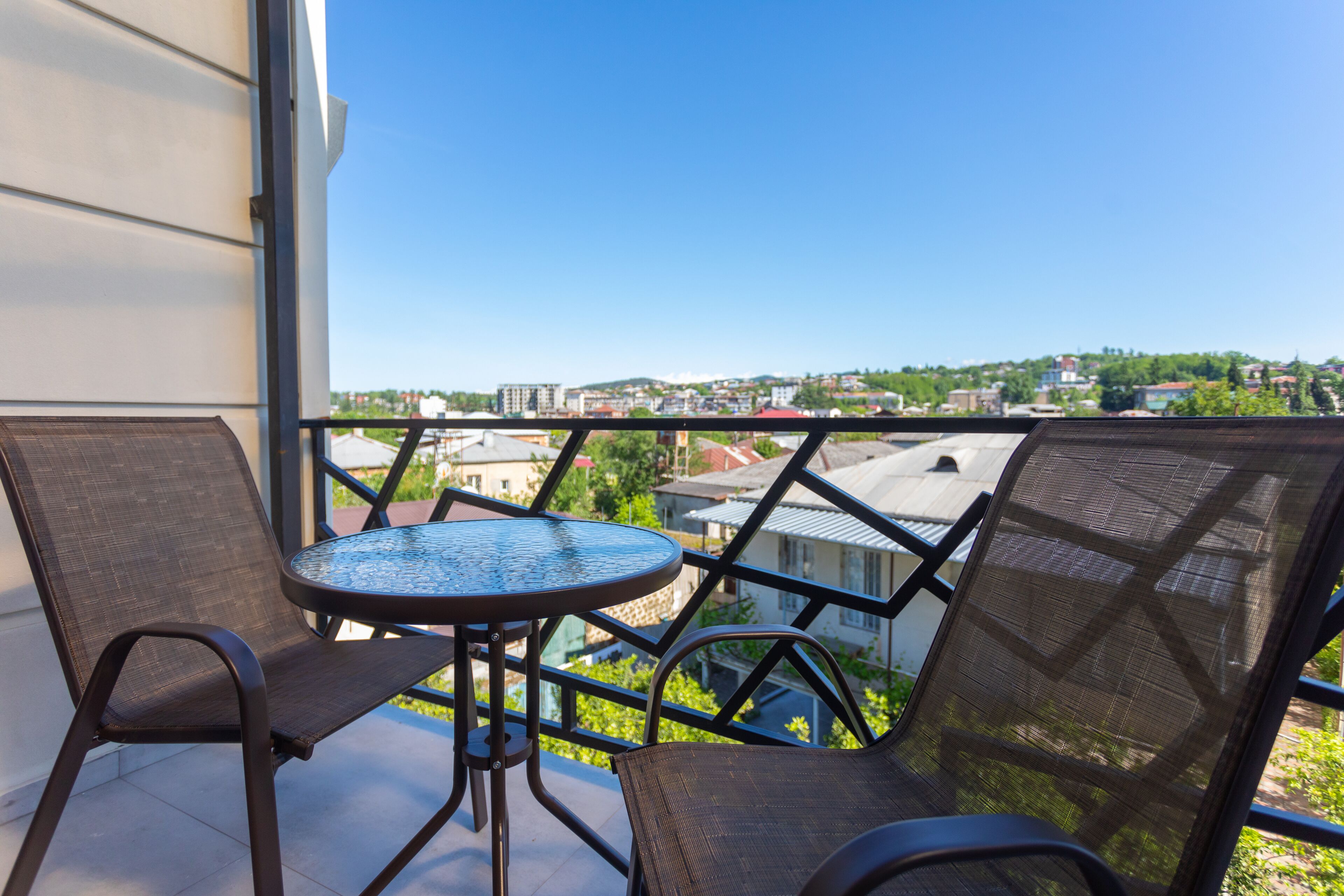 A balcony with two chairs and a small table, overlooking a cityscape with buildings and greenery.