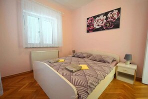 A bedroom with a bed, two bedside tables, a window with a white curtain, and a floral painting on the wall.
