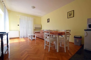 A room with a dining table, chairs, a sofa, a window with curtains, a wall-mounted air conditioner, and a trash bin.