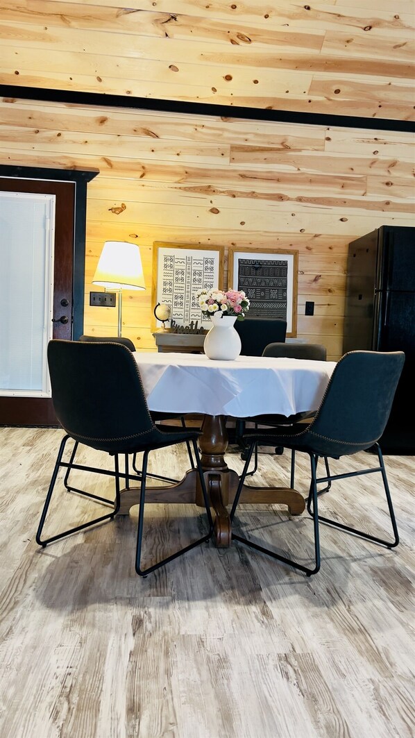 A dining area with a wooden ceiling, a table with a white tablecloth, two chairs, a vase with flowers, and two framed pictures on the wall. - Book 3 and Get 4th Night FREE! The Custard Apple Luxury Cabin with Hot Tub (Broken Bow)
