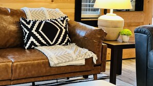 Un canapé en cuir marron agrémenté d’un coussin à motifs noir et blanc et d’un plaid blanc en tricot. Une table avec une lampe et une petite plante en pot se trouve devant le canapé.