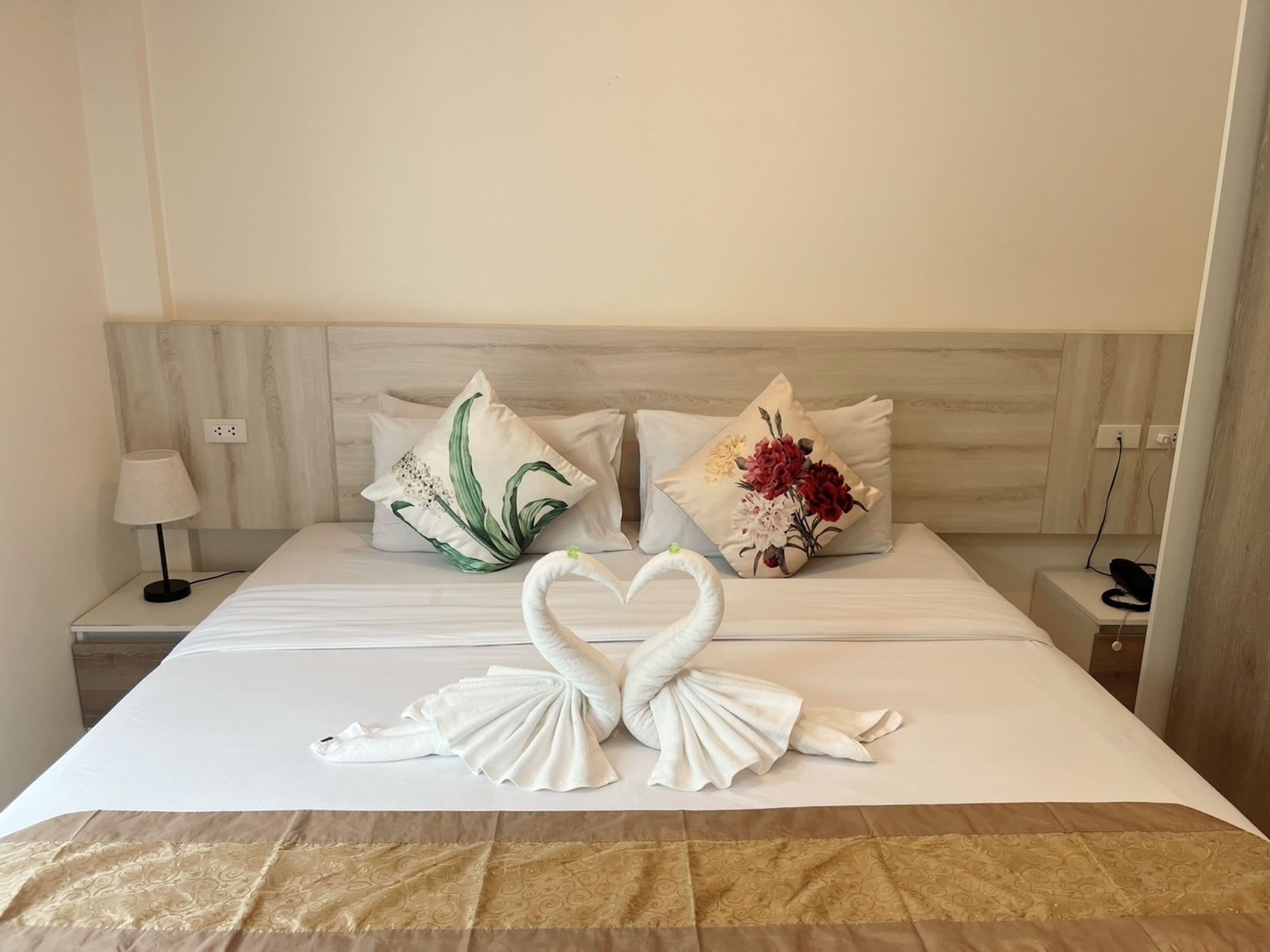 A hotel room with a neatly made bed, two pillows with floral designs, a lamp on the nightstand, and a swan-shaped towel arrangement on the bed.