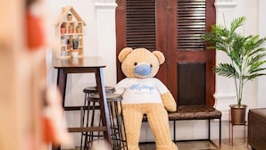 A plush teddy bear sitting on a bench in a cozy room with a wooden table, a small shelf with potted plants, and a potted palm plant. - Mystays Phuket (Ko Kaeo)