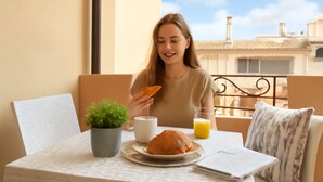 A person sitting at a table with a croissant, a cup, and a glass of orange juice. There's a small plant on the table and a view of buildings in the background. - Elegant two-room apartment with wifi, air conditioning, central area (Sardegna)