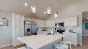 Una cocina moderna con armarios blancos, una isla grande con taburetes de bar, electrodomésticos de acero inoxidable e iluminación colgante.