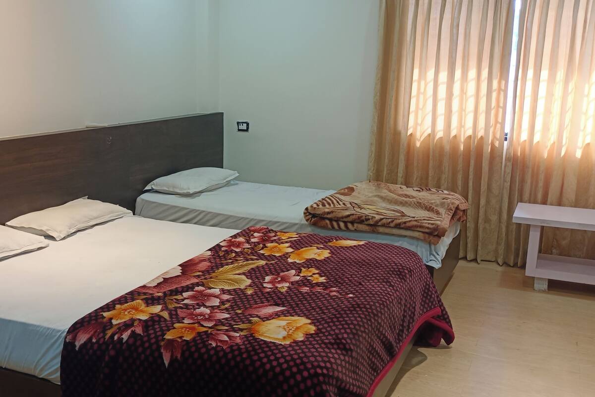Una habitación con dos camas, una con cubrecama floral y la otra con una cubierta lisa. Hay un pequeño banco blanco y una ventana con cortinas.