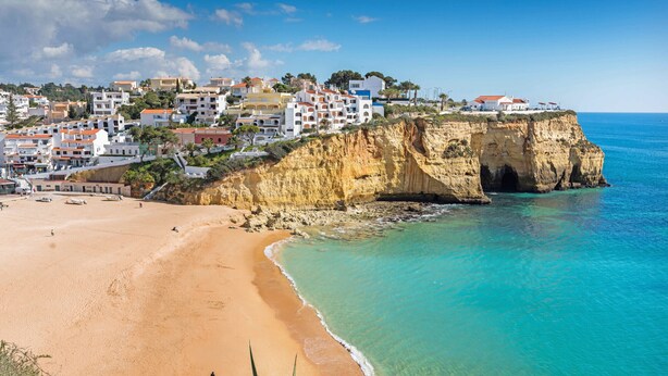 Un pueblo costero con edificios sobre un acantilado, con vista a una playa arenosa y aguas turquesas.