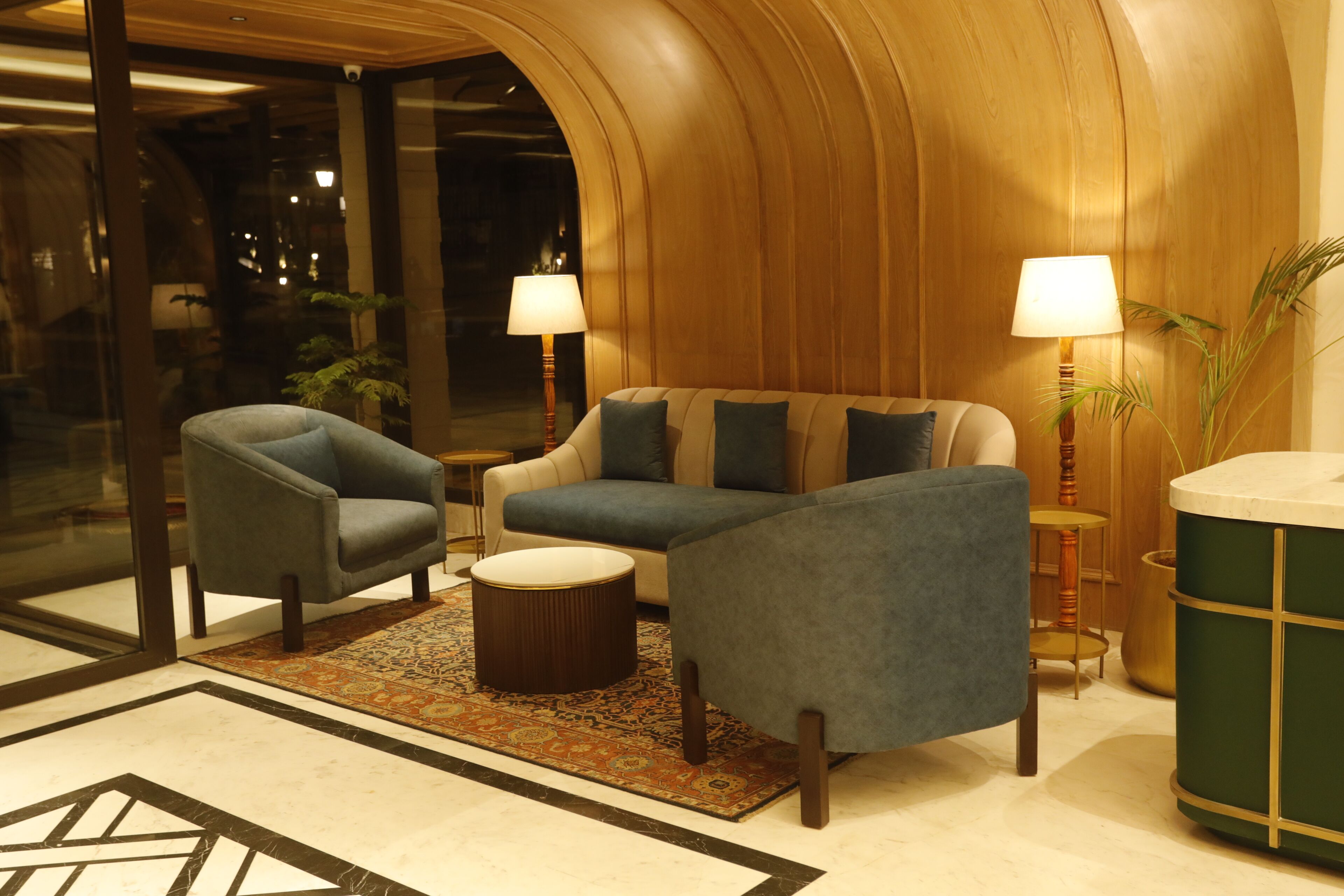 Un hall d’entrée moderne avec un mur en bois aux lignes courbes, un canapé bleu, deux fauteuils, un tapis à motifs et un pouf rond.
