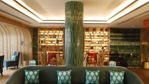 Um lobby moderno com um sofá verde, uma coluna cilíndrica alta e estantes com livros e objetos decorativos.