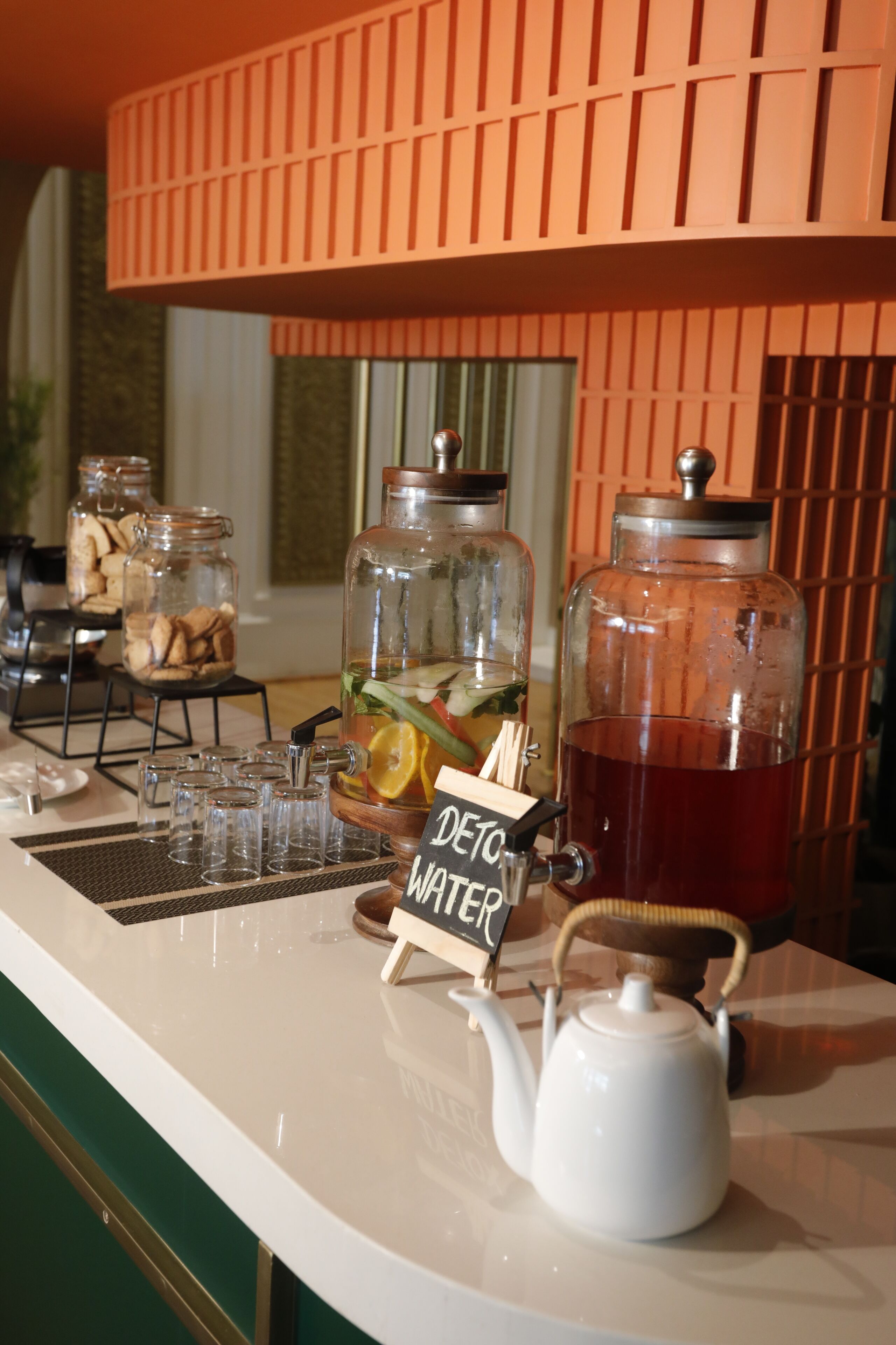 Una estación de bebidas con un letrero que dice 'DETO WATER', una tetera blanca y dos frascos de vidrio con comida.