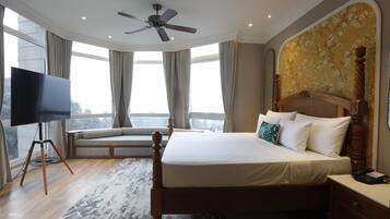 Habitación de hotel con una cama grande, un televisor sobre un atril, una mesita de noche de madera y vistas a la ciudad a través de amplios ventanales.