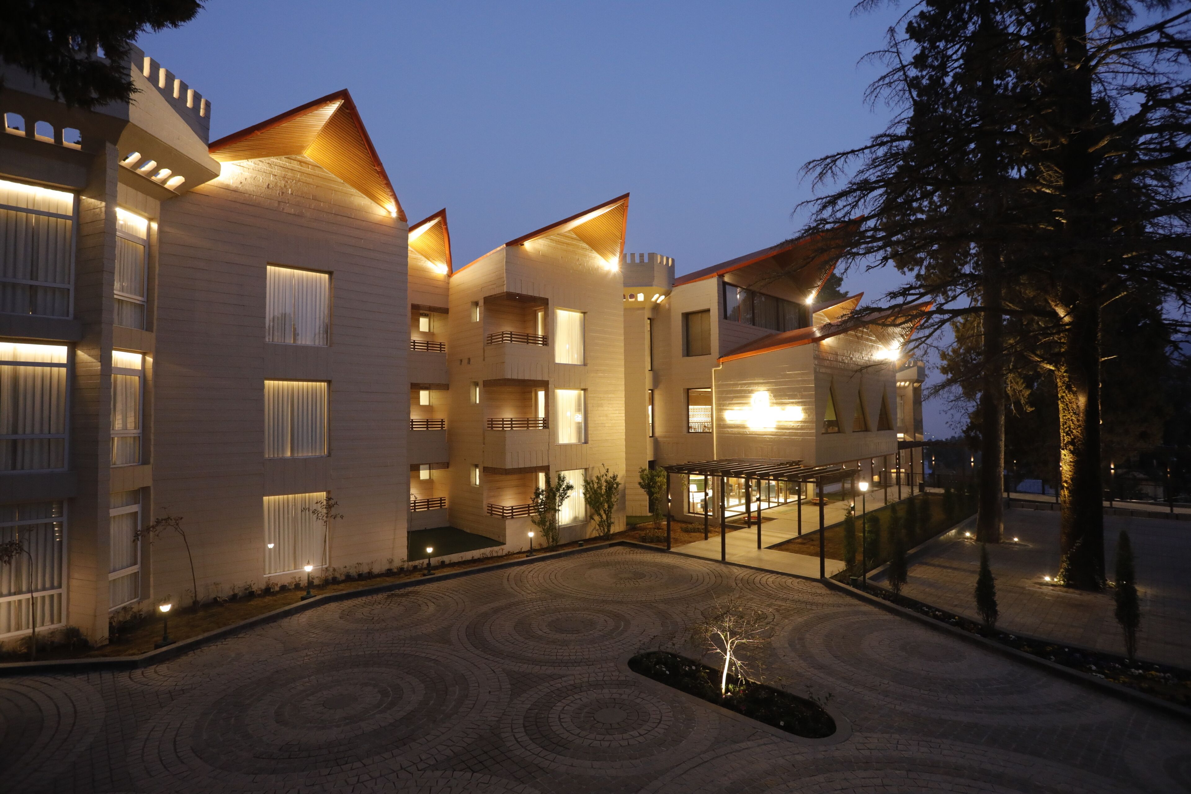Un hotel de varios pisos con balcones, un patio con pavimento estampado y una entrada bien iluminada.