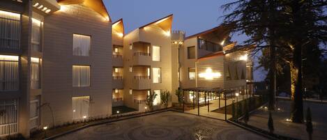 Un hotel de varios pisos con balcones, un patio con pavimento estampado y una entrada bien iluminada.