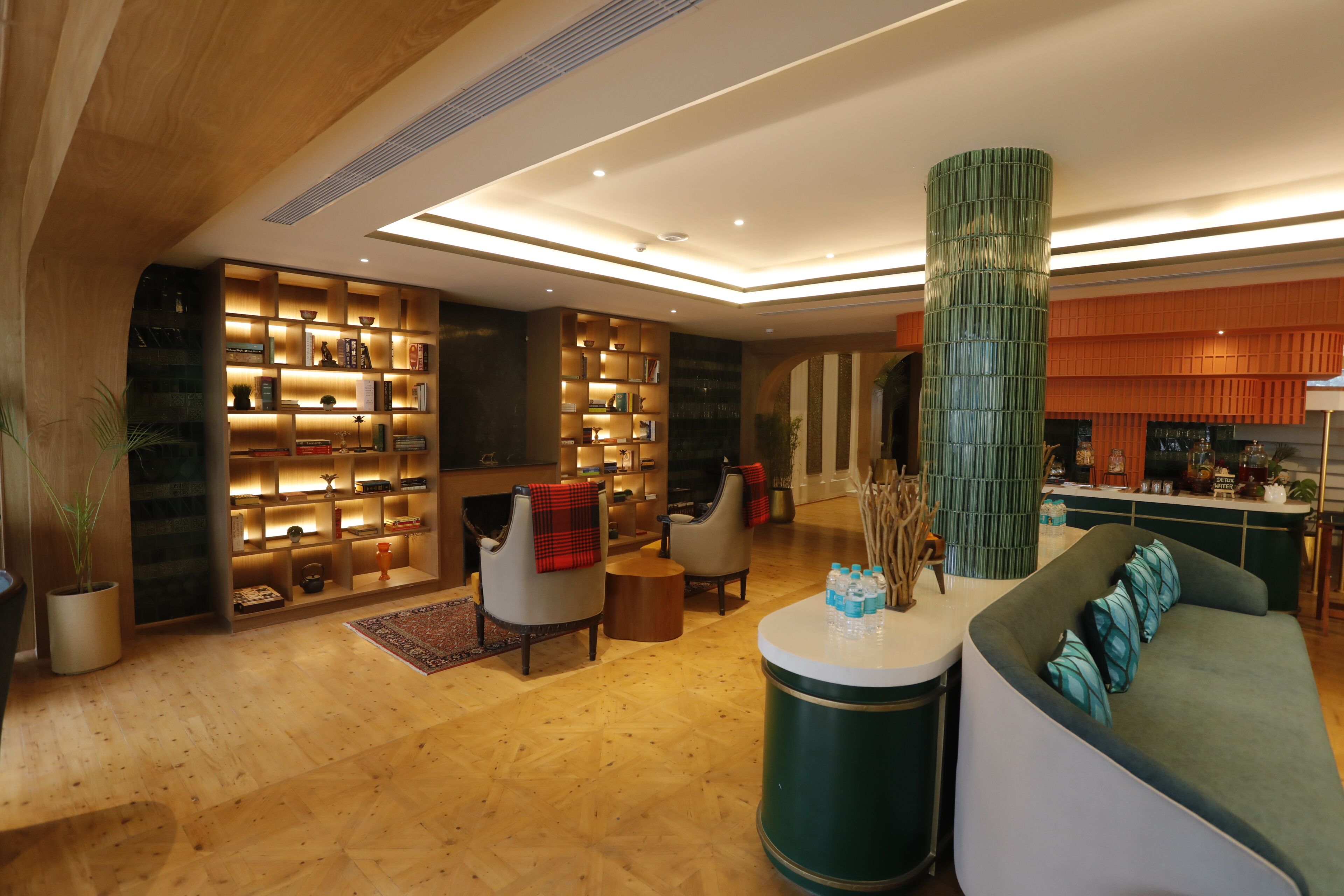 A modern lobby with a wooden floor, a green cylindrical pillar, a sofa with teal cushions, and a bar area.