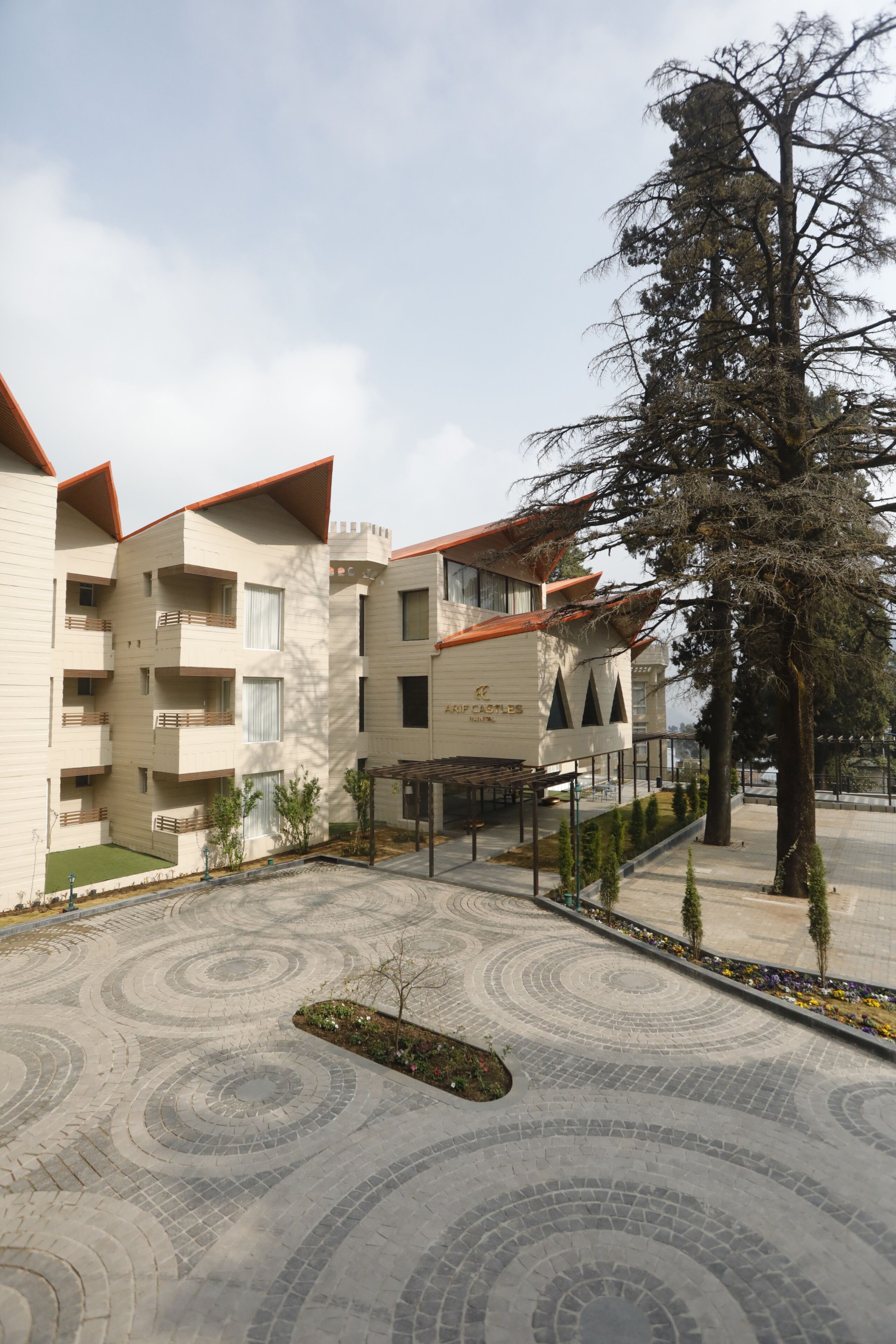 A multi-story building with balconies, a courtyard with patterned paving, and a large tree.