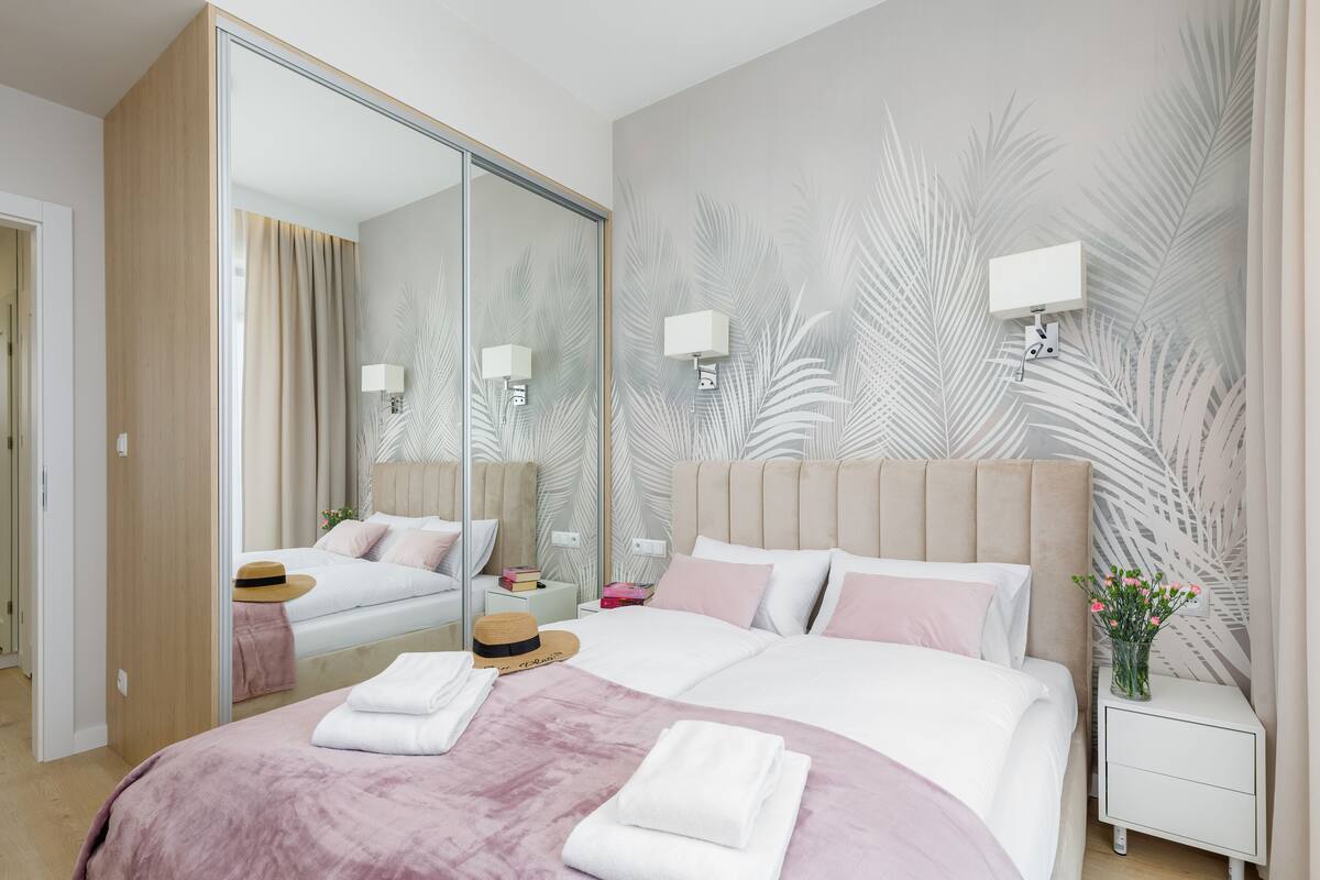 A modern hotel room with a large bed, a mirror, a nightstand with a lamp, and a wall with a leaf pattern.